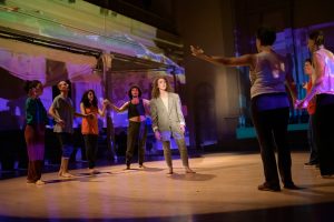 Dancers on a stage with an unclear image projected on their bodies and the stage, in two semi-circles, linked by their hands held with arms raised at their sides. The dancers are encircling the choreographer and performer as she is standing in the middle with her mouth agape and a lavelier microphone resting on her cheek. She is wearing an oversized gray suit with the ends of her sleeves rolled up.