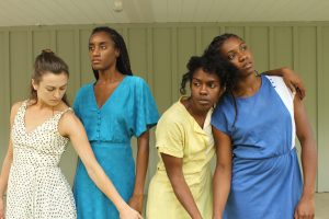 Four women in daylight, one woman face looking back reaches an arm behind her, the second stands staring into the distance, and the third has her arm draped over the fourth woman's shoulder.
